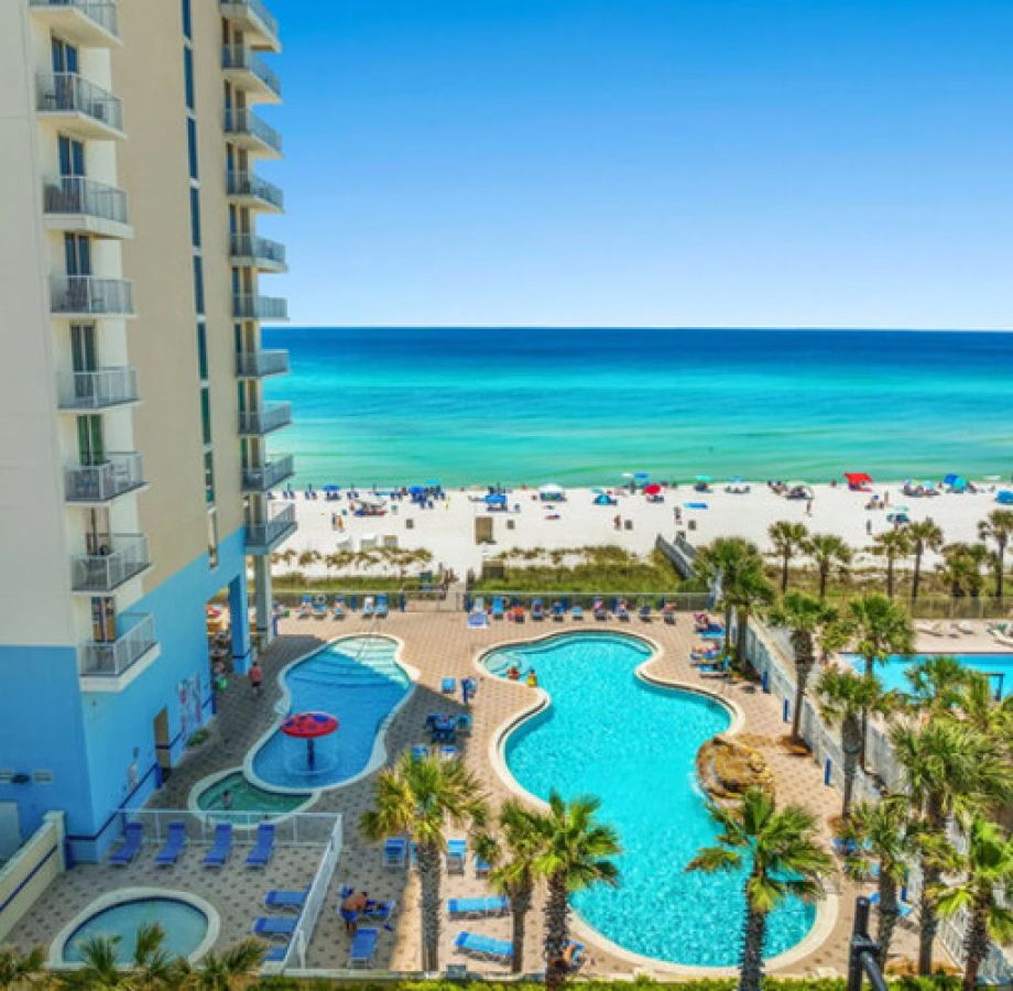 Majestic beach resort with oceanfront swimming pools, palm trees, white sand beach, and turquoise water under a clear blue sky.