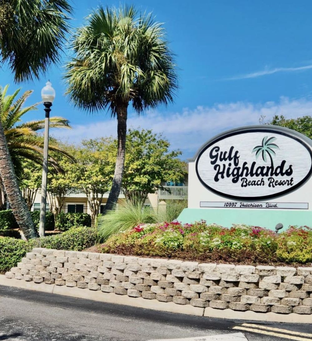 Front entrance sign for Gulf Highlands Beach Resort surrounded by palm trees and landscaped greenery.